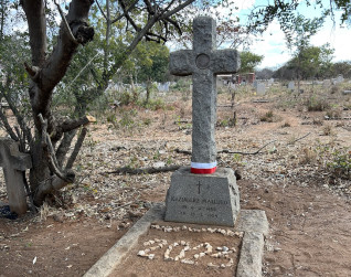 Grób Kazimierza Malujdy z kamiennym krzyżem i wstążką z polską flagą na cmentarzu w Livingstone, Zambia. Grób otoczony suchą ziemią i rzadką roślinnością.