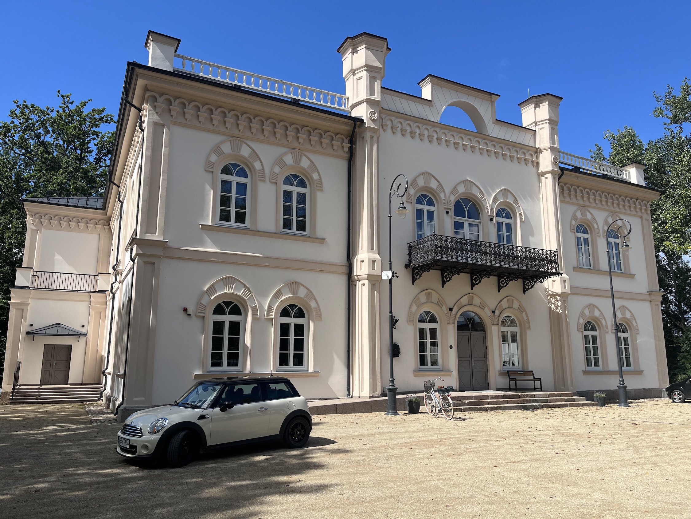 Le palais néogothique de Glinciszki avec sa façade lumineuse, ses fenêtres cintrées et son balcon décoratif. Une voiture et un vélo sont garés devant le bâtiment. Des arbres sont visibles à l'arrière-plan.