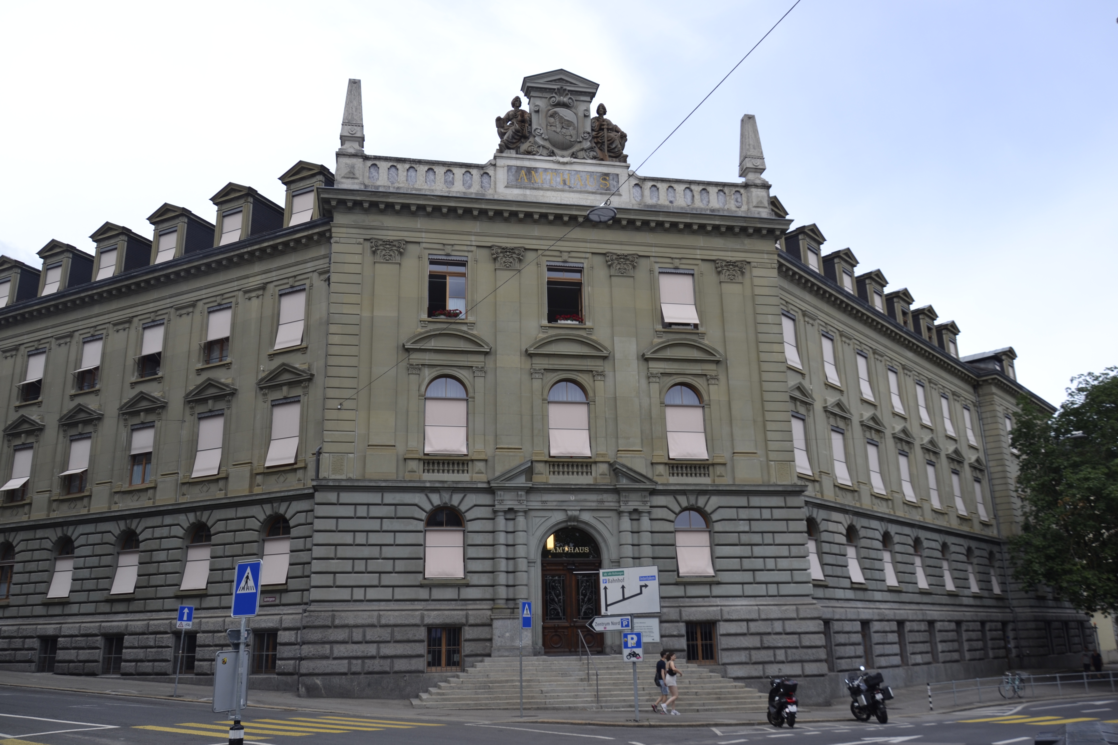 Le bâtiment Amthaus à Berne, conçu par l'architecte Franciszek Stempkowski, présente une façade néoclassique avec des fenêtres cintrées et un fronton décoratif. Deux motos sont garées devant.