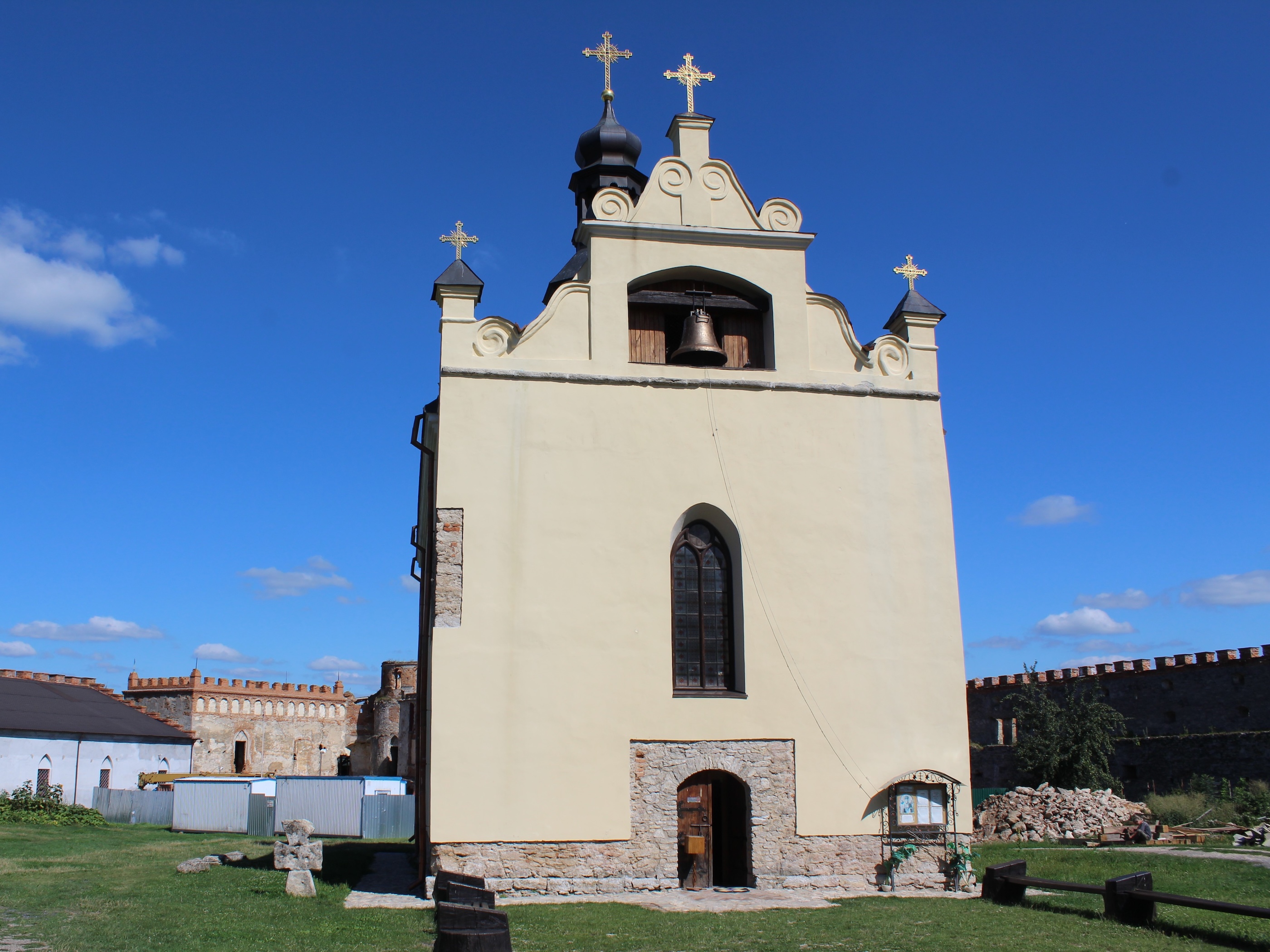 Une chapelle avec une cloche et une croix au sommet, faisant partie du complexe du château de Medzhybizh en Ukraine. Le bâtiment a une façade claire avec des fenêtres cintrées et une base en pierre. Ciel bleu clair en arrière-plan.