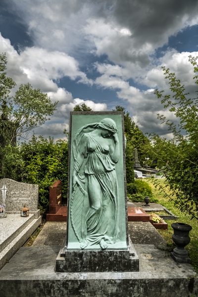 Gravestone of Józef Wieniawski at Ixelles Cemetery in Brussels, featuring a bronze relief of Euterpe, the Muse of Music, by Hendrik Joos. The gravestone is surrounded by greenery and other tombstones.