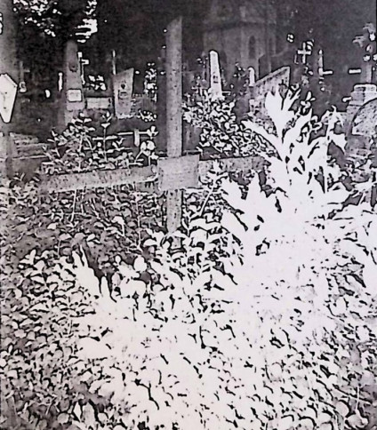 Krzyż żeliwny z rurek oznacza grób, na przecięciu ramion tabliczka ołowiana z inskrypcją. Widoczna otaczająca roślinność.