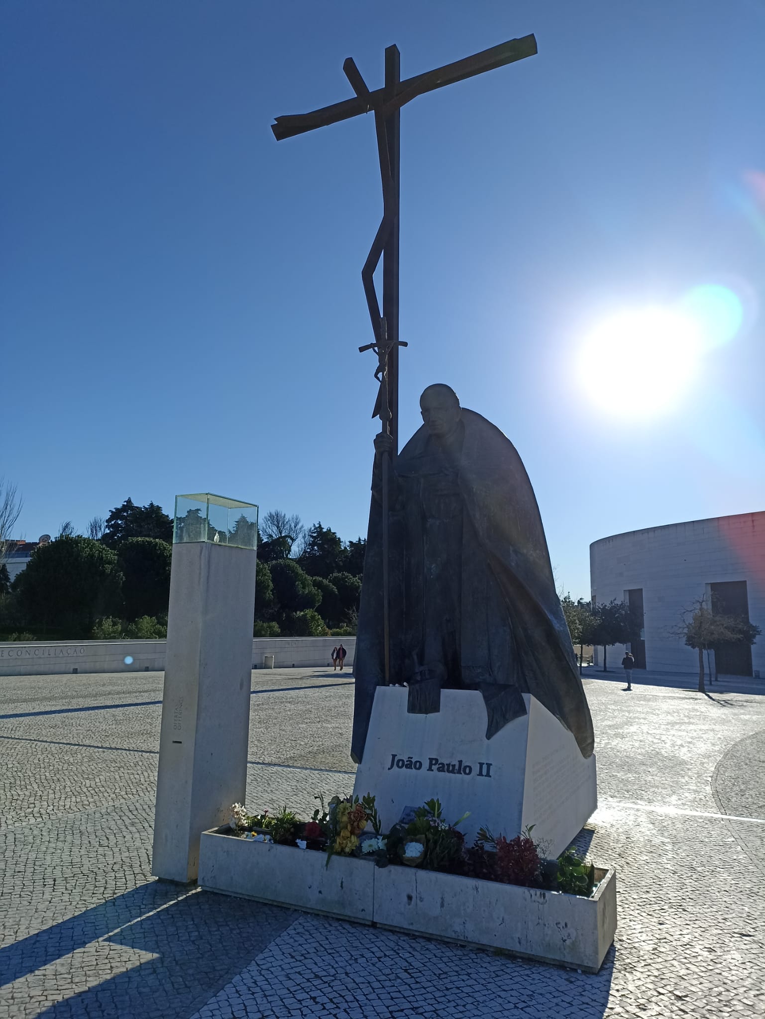 Statue de Jean-Paul II tenant une grande croix, avec des fleurs à la base, sous un ciel bleu clair. Le soleil brille intensément en arrière-plan.