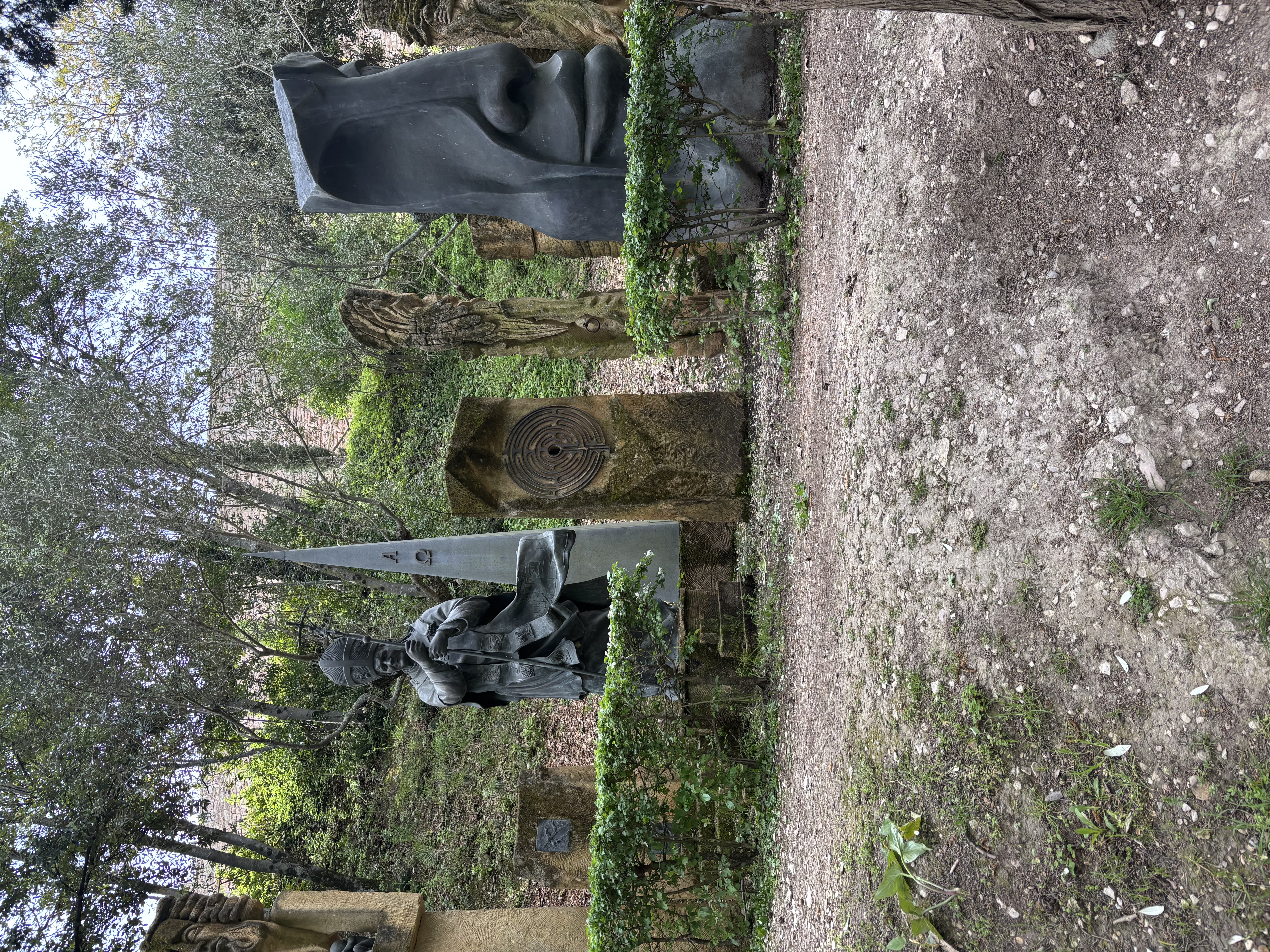 Sculpture de Jean-Paul II tenant une pastorale, entourée de sculptures abstraites en pierre et en métal dans une zone boisée. Verdure et chemin de terre en arrière-plan.