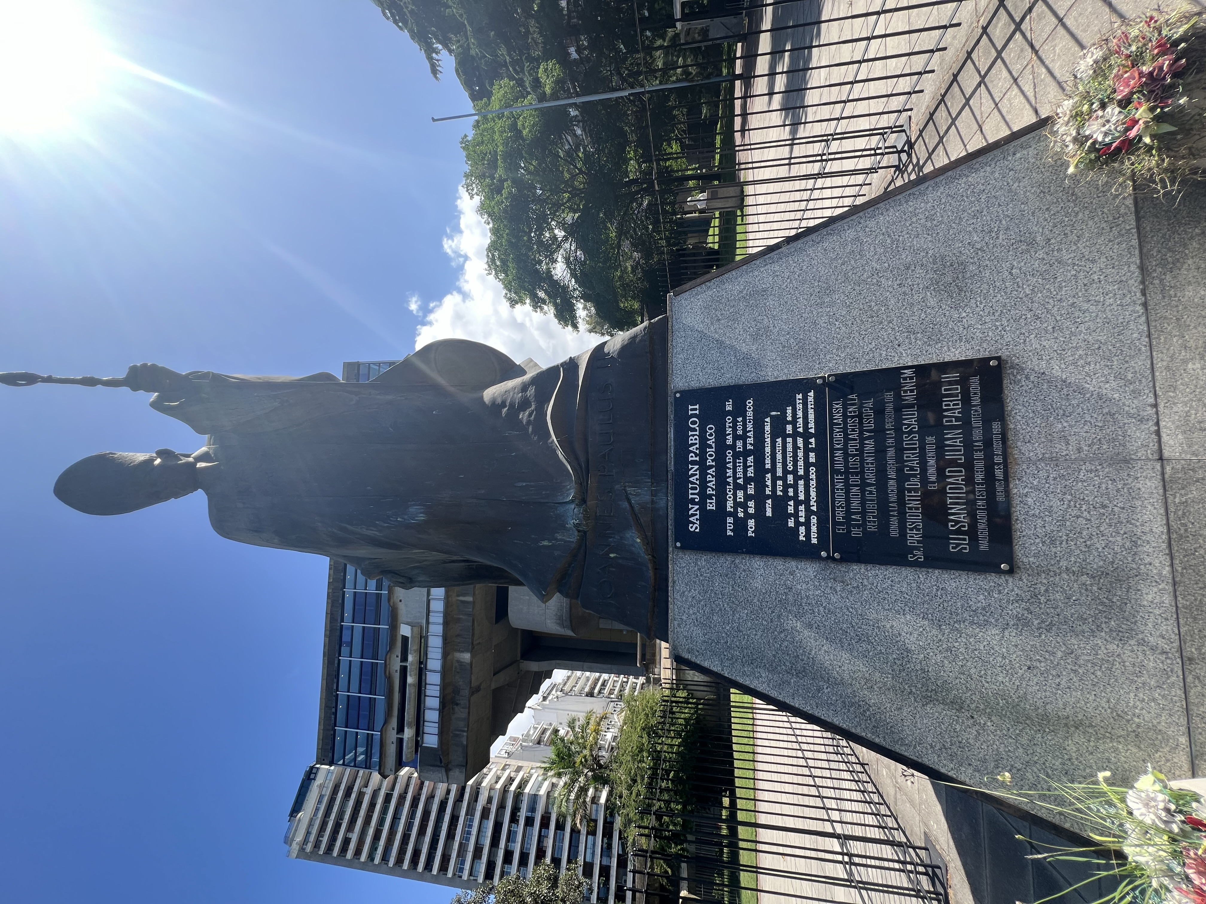 Bronze statue of John Paul II in front of a modern building, with a plaque in Spanish describing his canonisation and services. Sunlight falls from the upper right corner.