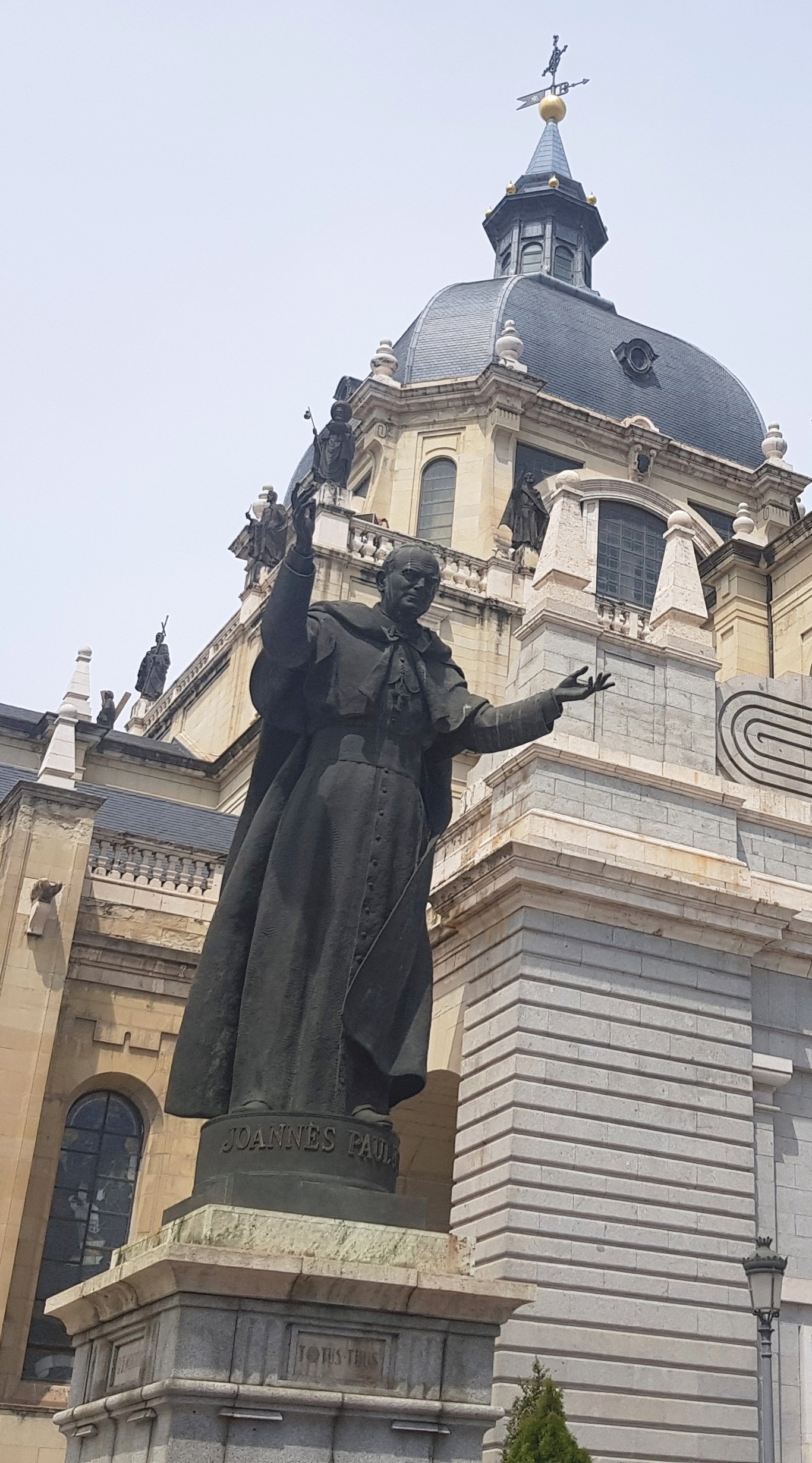 Statue de Jean-Paul II, bras tendus, devant une grande cathédrale avec dôme et flèche. Le socle de la statue porte l'inscription "Joannes Paulus II".