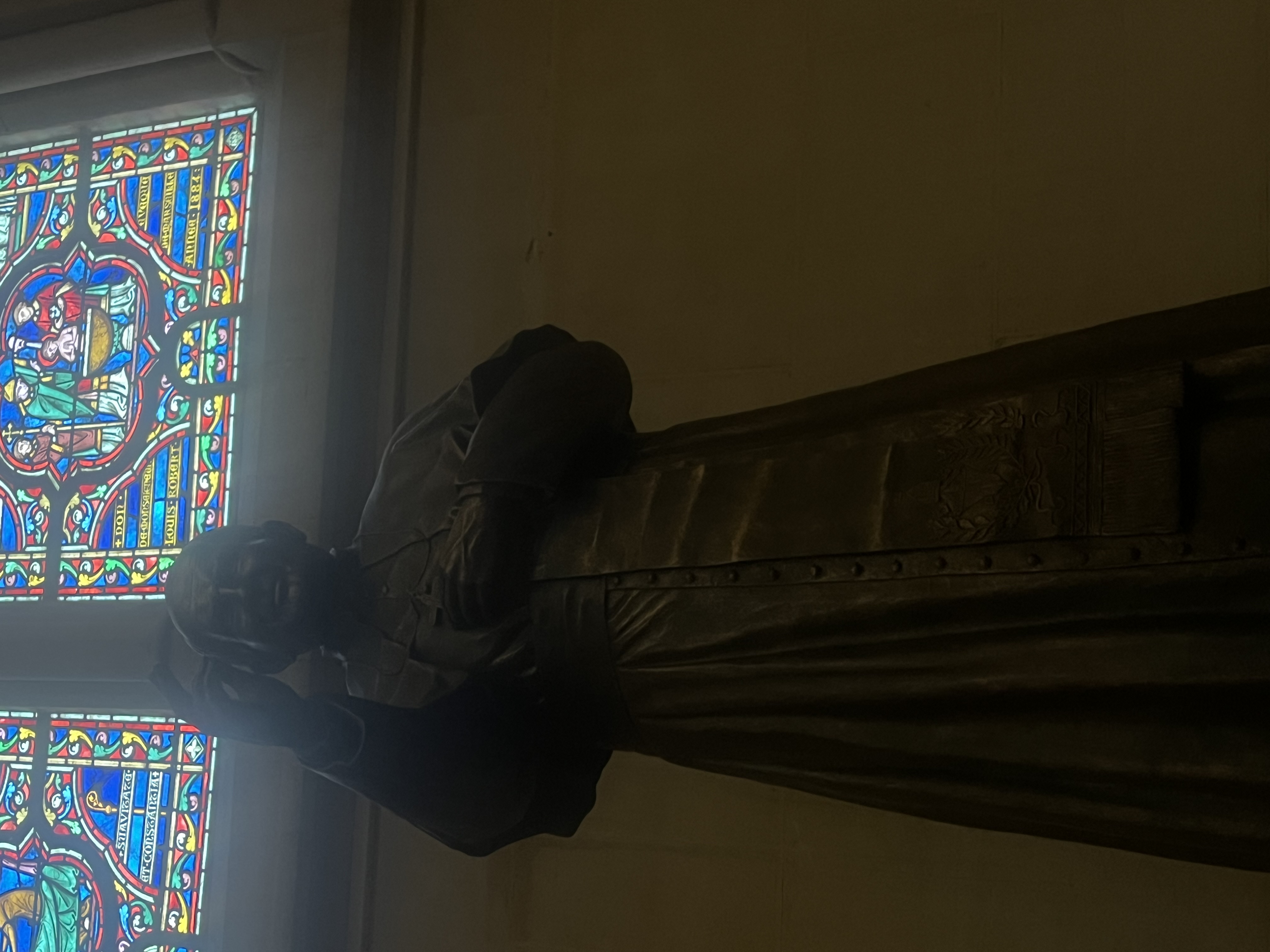 Statue en bronze du pape Jean-Paul II à l'intérieur d'une église, avec un vitrail coloré au-dessus représentant des figures religieuses.
