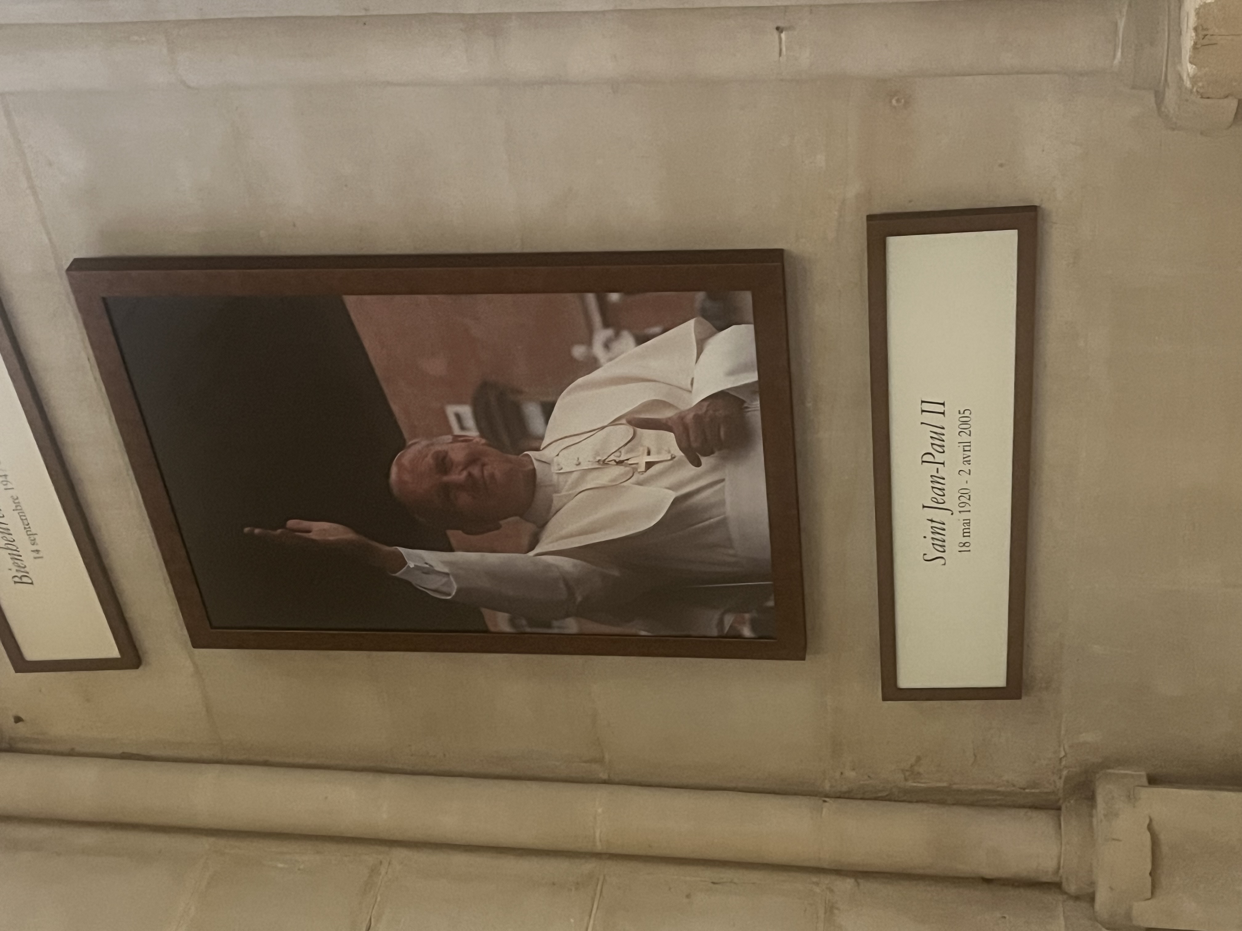 Photographie encadrée de Jean-Paul II saluant, avec une plaque en dessous indiquant 'Saint Jean-Paul II, 18 mai 1920 - 2 avril 2005' sur un mur en pierre.