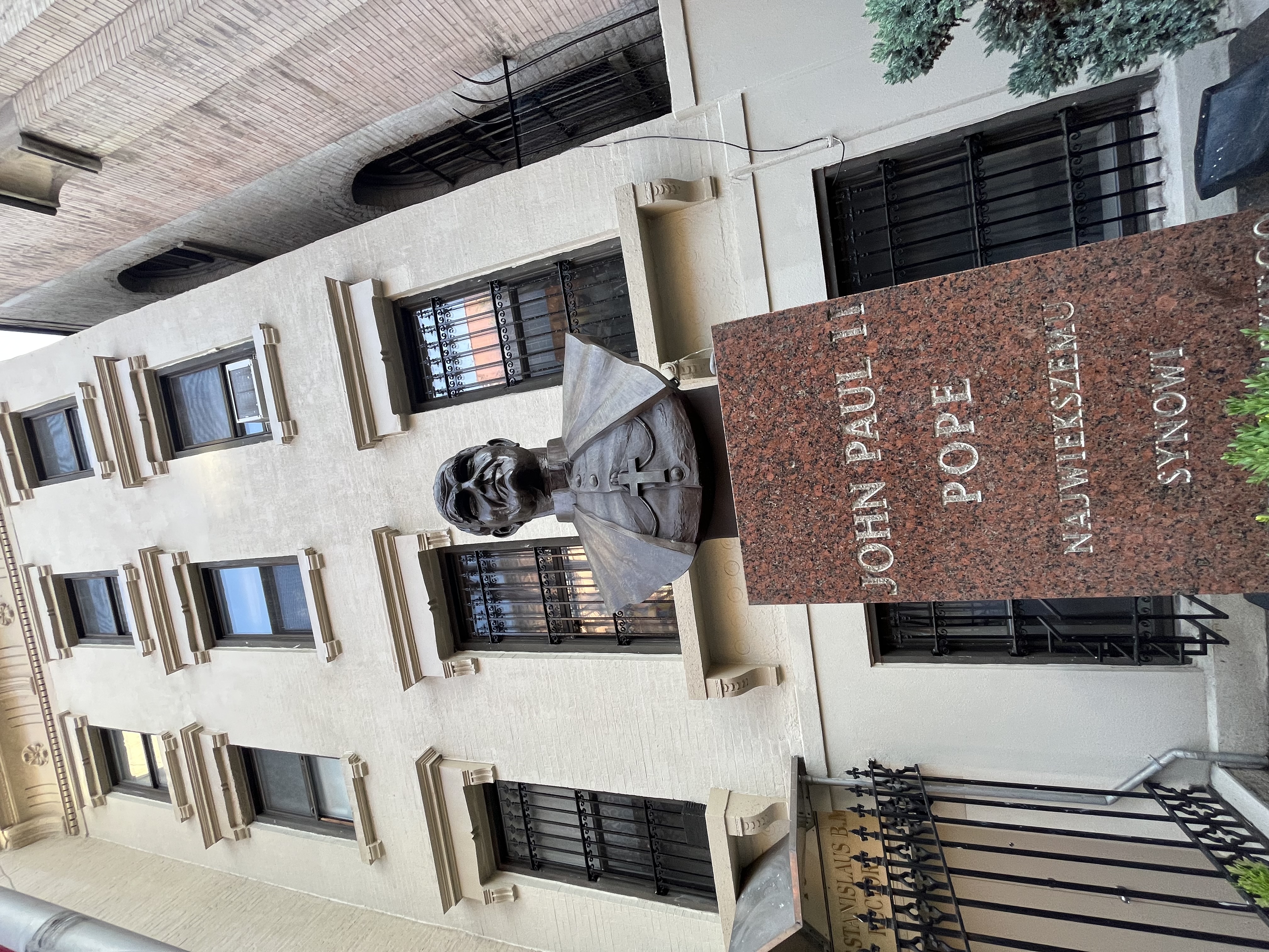 Bronze bust of John Paul II on a red granite pedestal with inscriptions in Polish and English, in front of a beige building with barred windows.