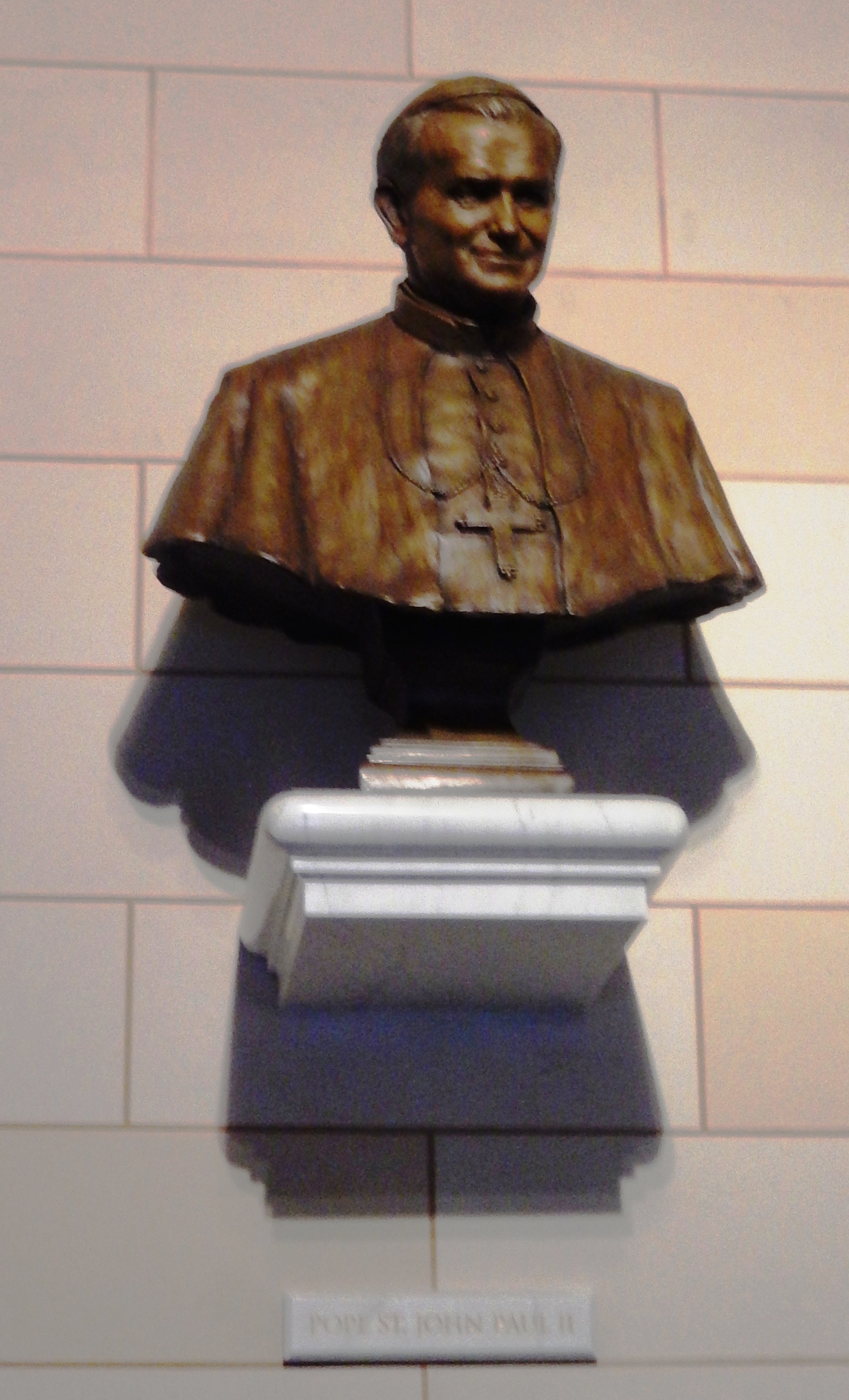 Buste en bronze du pape Jean-Paul II sur un socle blanc contre un mur carrelé. La plaque en dessous indique 'Pape St. Jean-Paul II'.