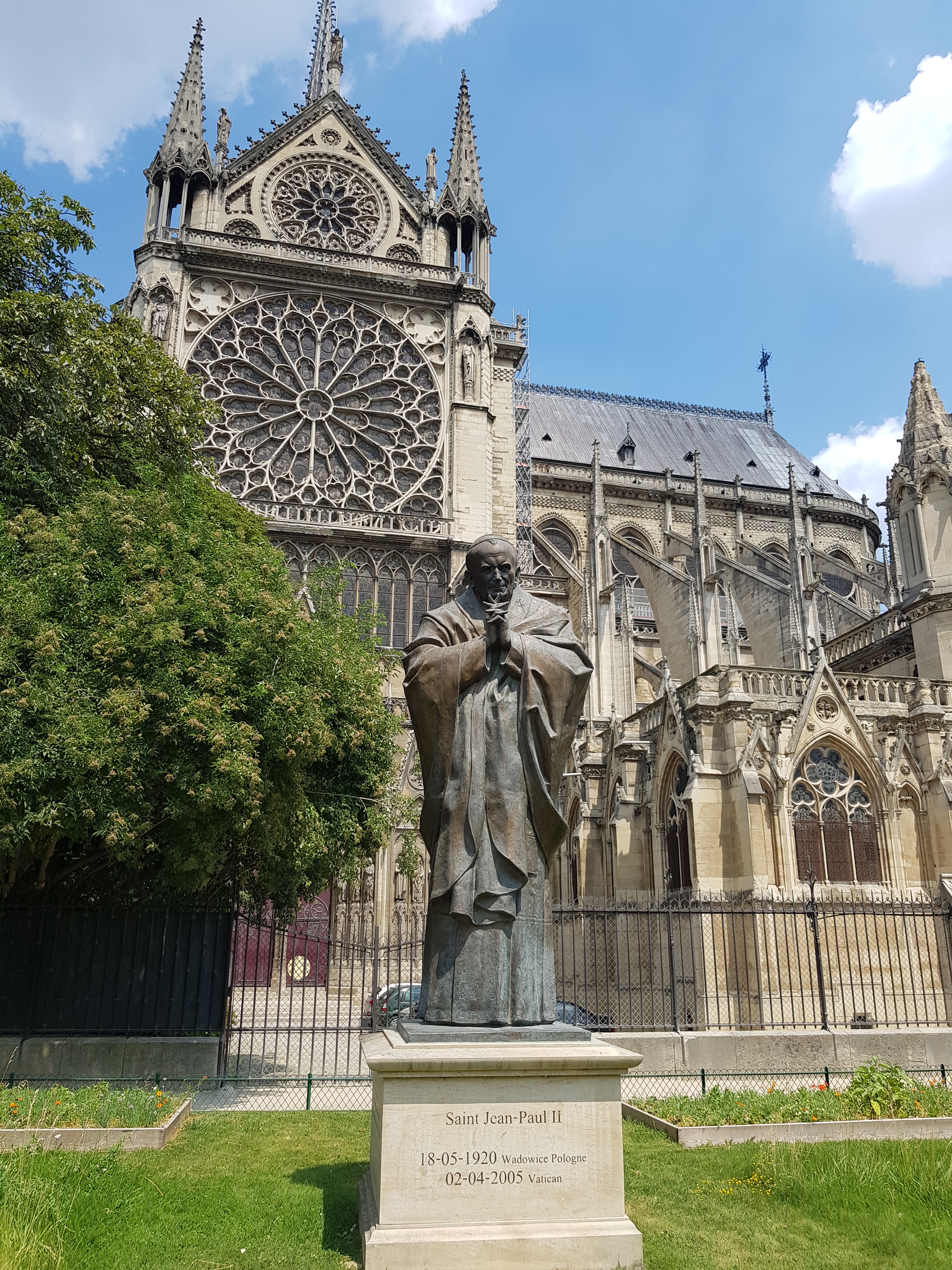 Statue de Saint Jean-Paul II devant la cathédrale Notre-Dame de Paris. La statue est sur un piédestal avec les inscriptions de ses dates de naissance et de décès. L'architecture gothique de la cathédrale est visible en arrière-plan.