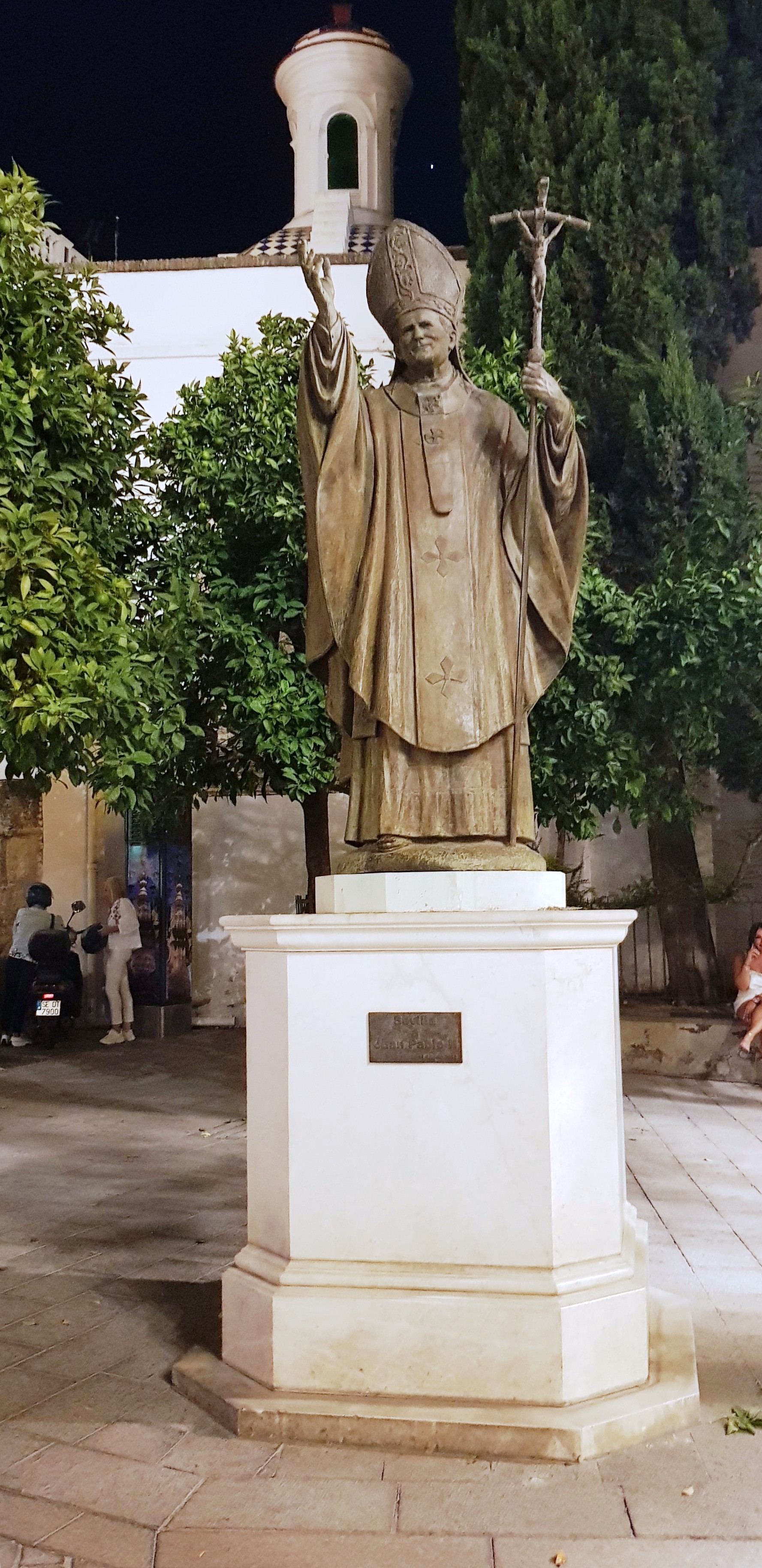 Statue en bronze du pape Jean-Paul II sur un piédestal, une main levée en bénédiction et tenant un bâton surmonté d'une croix. Située dans une cour avec des arbres et un bâtiment en arrière-plan.