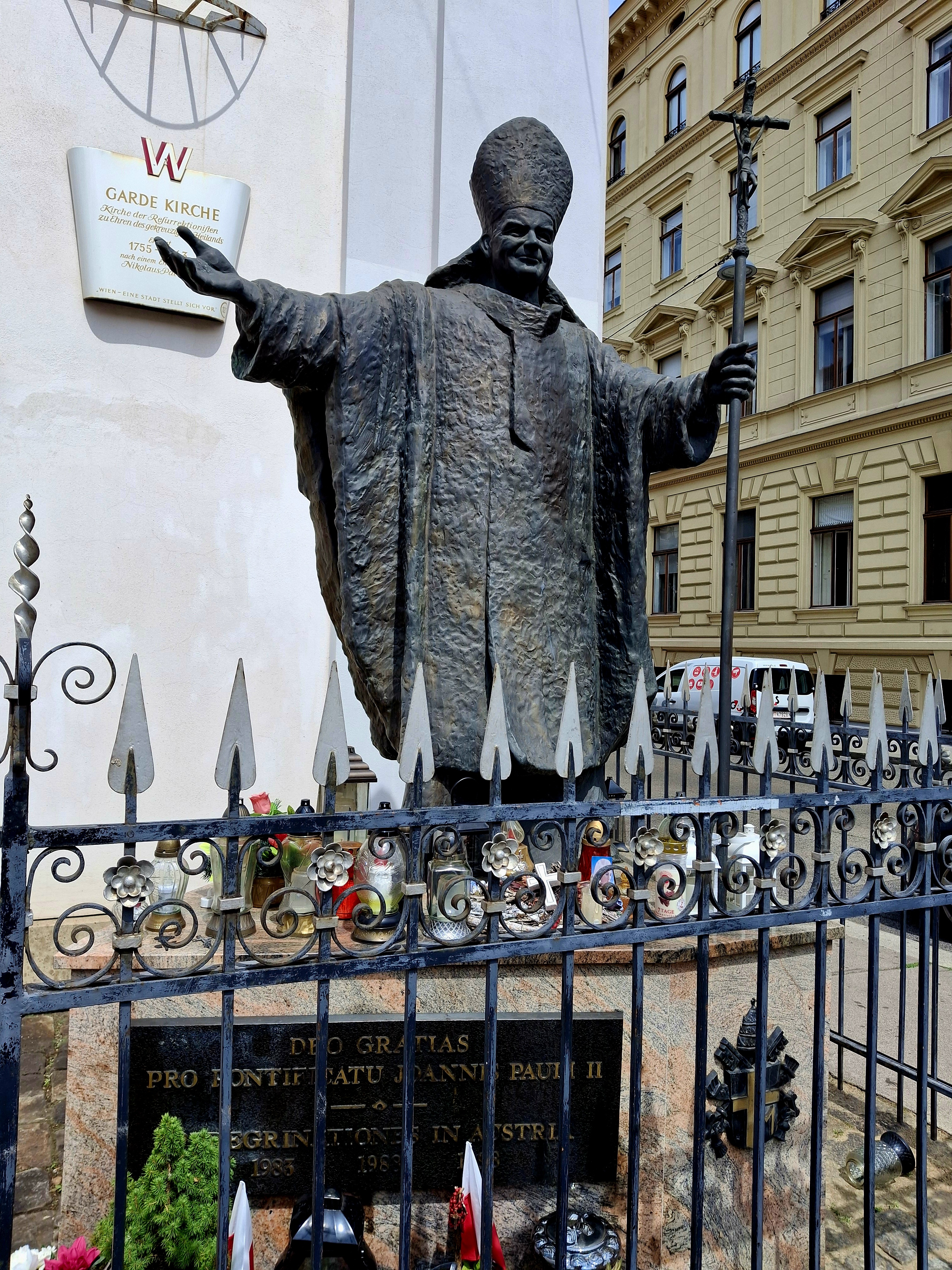 Statue en bronze du pape Jean-Paul II avec les bras étendus, tenant un crucifix, derrière une clôture en fer décorative. Une plaque avec du texte en latin est visible à la base.