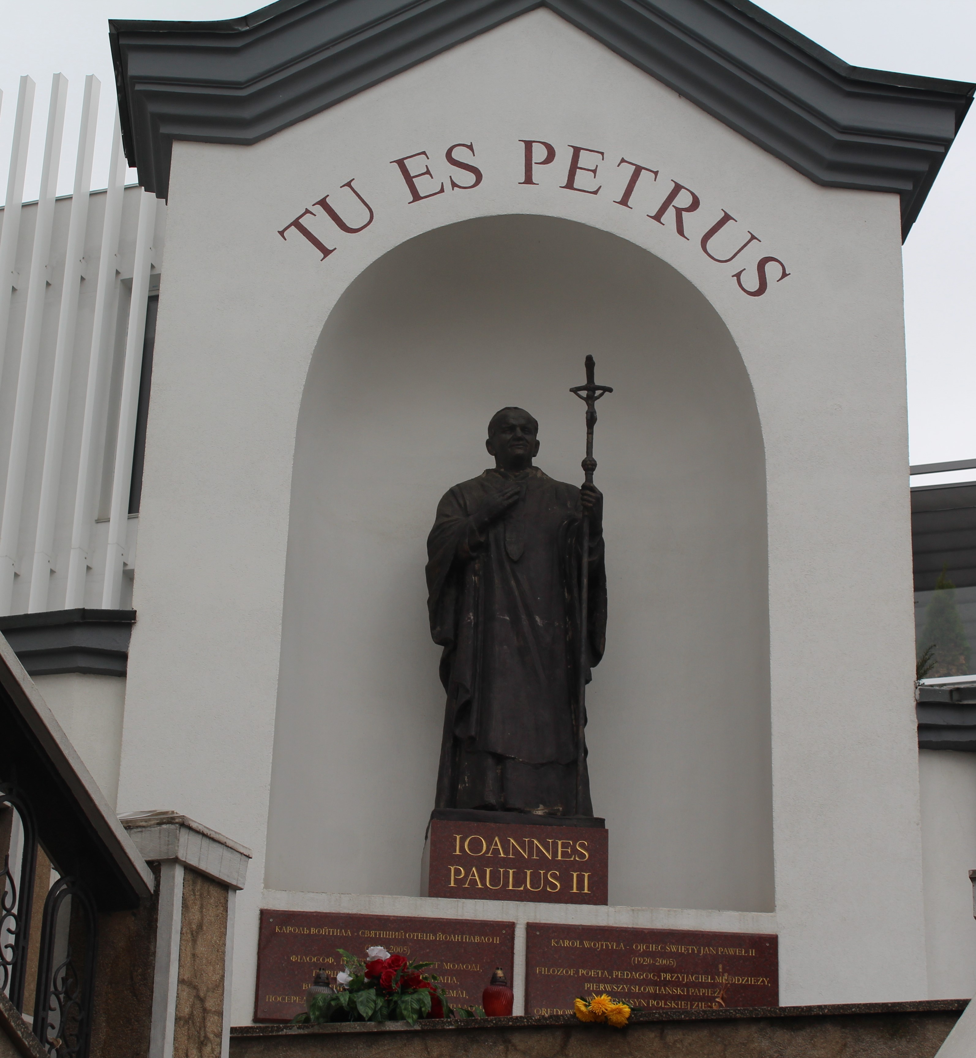 Statue de Jean-Paul II tenant une croix, placée dans une alcôve avec l'inscription 'TU ES PETRUS' au-dessus. En dessous, des plaques avec du texte en plusieurs langues et des fleurs.