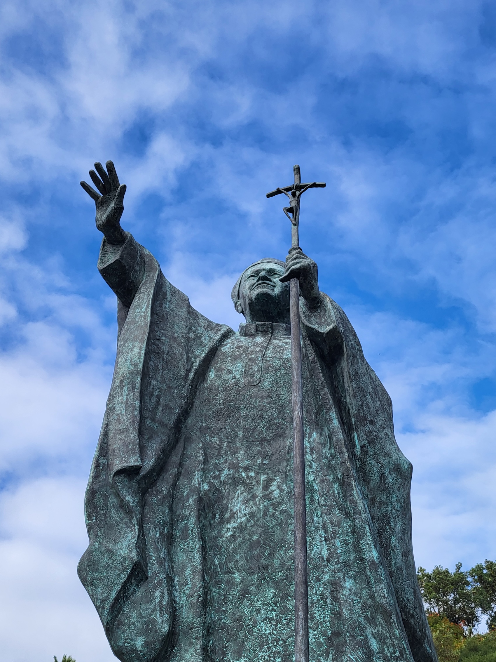 Statue en bronze du pape Jean-Paul II avec la main levée et tenant un crucifix, sur fond de ciel bleu avec des nuages.