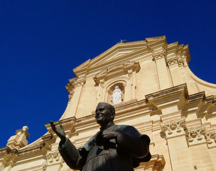 Statue of John Paul II in front of a church with a blue sky background. The church facade is detailed with a niche containing a statue.
