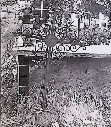 Żeliwny krzyż oznaczający grób Alberta Zdzijowskiego, z ozdobnymi wolutami i tabliczką u podstawy. Krzyż ma spiralne i wolutowe zdobienia, na tle cmentarza.