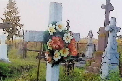 Nagrobek z krzyżem na cokole, z płyciną z inskrypcją. Podstawa ma ścięte górne krawędzie. Krzyż ozdobiony wieńcem kwiatowym. Wokół inne nagrobki na trawiastym terenie.