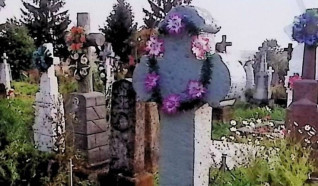 Grób oznaczony krzyżem z ramionami zakończonymi trójliściem na cokole. Cokół ma ścięte górne krawędzie i rytą inskrypcję z przodu. Widoczne otaczające groby i kwiaty.