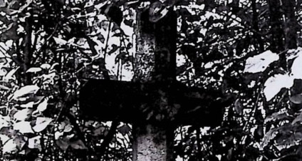 Nagrobek Yaniny Lipińskiej z krzyżem na zwężającym się cokole, o przekroju ośmioboku i wieńcem na przecięciu ramion, otoczony liśćmi.