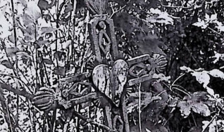 Żeliwny krzyż na cokole z ramionami zakończonymi palmetami i tablicą w kształcie serca na przecięciu. Krzyż zdobiony rombami i esownicami, otoczony roślinnością.