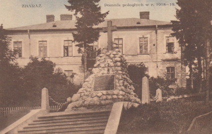Pocztówka przedstawiająca pomnik w Zbarażu z lat 30. XX wieku, upamiętniający poległych z lat 1918-1919. Kamienna konstrukcja z krzyżem i tablicą znajduje się przed budynkiem.