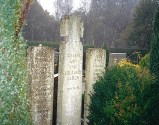 Nagrobki na cmentarzu w Langholt, w tym centralna tablica z piaskowca z krzyżem Wehrmachtu i cytatem z Jana 14:19. Otaczająca zieleń i drzewa w tle.
