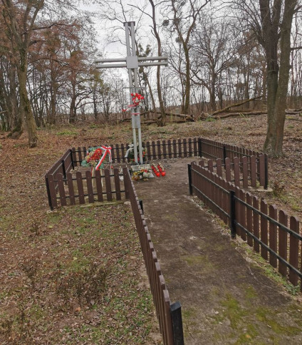 Metalowy krzyż z tabliczką stoi w ogrodzonej części cmentarza katolickiego, otoczony drzewami. U podstawy krzyża znajdują się kwiaty i wieńce.