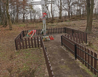 Metalowy krzyż z tabliczką stoi w ogrodzonej części cmentarza katolickiego, otoczony drzewami. U podstawy krzyża znajdują się kwiaty i wieńce.