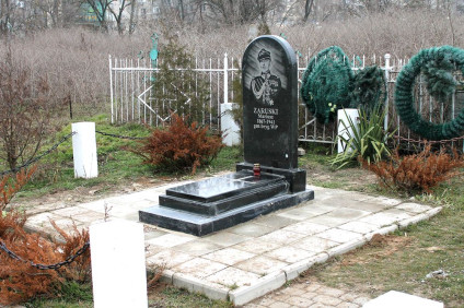 Grób Mariusza Zaruskiego z czarną płytą nagrobną z jego portretem i napisem, otoczony białym ogrodzeniem i zielenią.