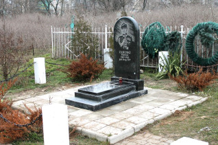 Grób Mariusza Zaruskiego z czarną płytą nagrobną z jego portretem i napisem, otoczony białym ogrodzeniem i zielenią.