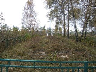 Trawiasty kopiec z metalowym krzyżem na szczycie, otoczony jasnoniebieskim metalowym ogrodzeniem. W tle stoją drzewa z jesiennymi liśćmi.