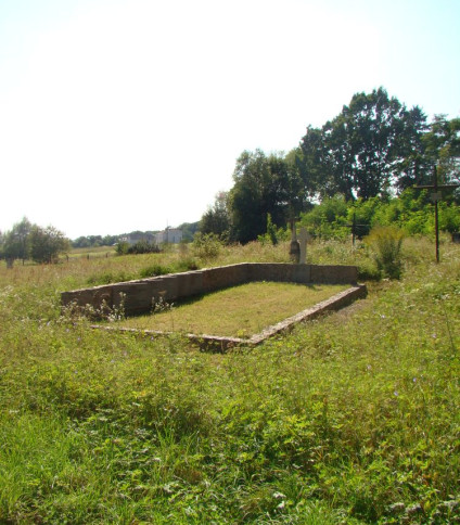 Grób ofiar bombardowania na stacji kolejowej Komarno-Buczały, wrzesień 1939. Kamienny krzyż stoi na czele grobu, otoczony zarośniętą trawą i drzewami w tle.
