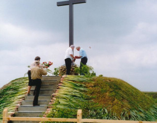 Symboliczny grób z dużym krzyżem upamiętniający żołnierzy 27. Wołyńskiej Dywizji Piechoty AK. Ludzie składają kwiaty na kopcu, otoczonym zielenią i drewnianym ogrodzeniem.