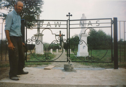 Mężczyzna stoi obok kutego żelaznego ogrodzenia z napisem 'ZBAW NAS' na cmentarzu. Za ogrodzeniem znajdują się krzyże i nagrobki otoczone zielenią.