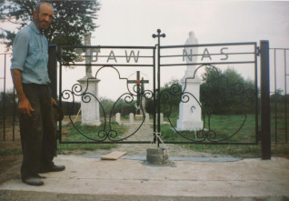 Mężczyzna stoi obok kutego żelaznego ogrodzenia z napisem 'ZBAW NAS' na cmentarzu. Za ogrodzeniem znajdują się krzyże i nagrobki otoczone zielenią.