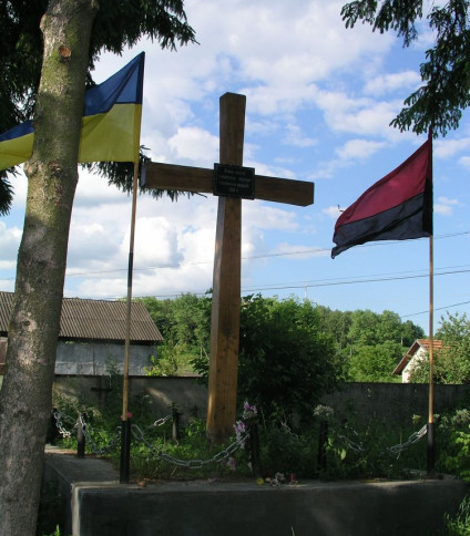 Drewniany krzyż na grobie z ukraińskimi flagami po obu stronach. Grób otoczony łańcuchem i kwiatami. Napis w języku ukraińskim upamiętnia ofiary represji stalinowskich z 1941 roku.