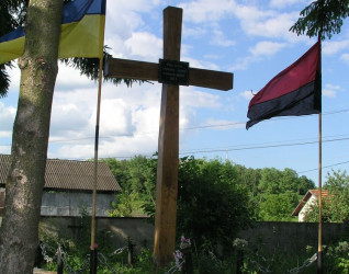Drewniany krzyż na grobie z ukraińskimi flagami po obu stronach. Grób otoczony łańcuchem i kwiatami. Napis w języku ukraińskim upamiętnia ofiary represji stalinowskich z 1941 roku.