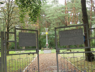 Brama wejściowa na cmentarz 27. Wołyńskiej Dywizji Piechoty Armii Krajowej. Widoczne są dwie tablice z napisami, a w tle wśród drzew stoi wysoki krzyż.