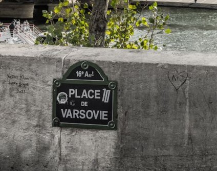 Widok na Wieżę Eiffla przez drzewa z Place de Varsovie w Paryżu, z tabliczką wskazującą lokalizację i Sekwaną w tle.