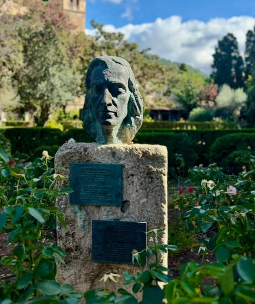 Bust of Fryderyk Chopin on a stone pedestal with plaques, surrounded by a garden with roses and trees in the background.
