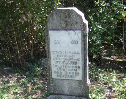 Nagrobek Ludwika Młokosiewicza z marmurową tablicą inskrypcyjną na prostokątnej podstawie z lastryko, otoczony zielenią. Tablica zawiera tekst w kilku językach, w tym daty 1831 i 1909.