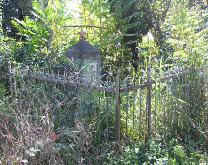Cementowy obelisk nagrobka Jerzego Sokołowskiego z czerwoną gwiazdą na szczycie, otoczony metalowym ogrodzeniem. Widoczna marmurowa tabliczka z rosyjską inskrypcją i zdjęciem zmarłego. Teren zarośnięty roślinnością.