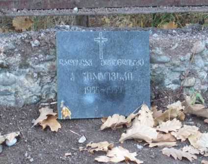 Nagrobek z czarnego marmuru z wyrytym krzyżem i inskrypcjami w obcym piśmie, otoczony opadłymi liśćmi i ziemią.