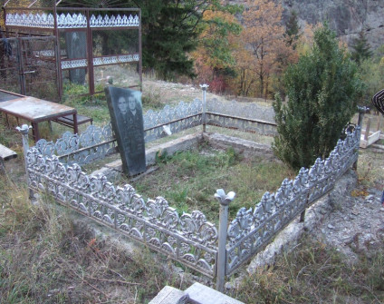 Nagrobek z czarnego granitu z wytrawionym wizerunkiem zmarłej, otoczony ozdobnym metalowym ogrodzeniem. Obok rośnie mały krzew, w tle drzewa i wzgórza.