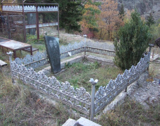 Nagrobek z czarnego granitu z wytrawionym wizerunkiem zmarłej, otoczony ozdobnym metalowym ogrodzeniem. Obok rośnie mały krzew, w tle drzewa i wzgórza.