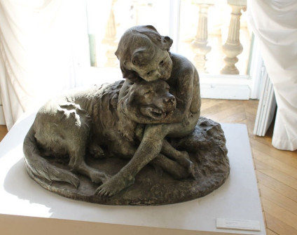 Brązowa rzeźba przedstawiająca dziecko obejmujące dużego psa, umieszczona na postumencie w pomieszczeniu z drewnianą podłogą i oknem w tle w Muzeum Sztuk Pięknych Julesa Chéreta w Nicei.