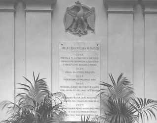 Tablica Biblioteki Polskiej w Paryżu z inskrypcjami o jej historii, otoczona dekoracyjnymi roślinami. Nad tablicą znajduje się emblemat orła.