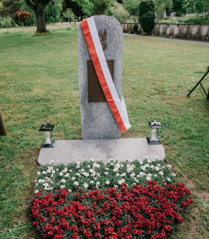 Granitowy obelisk z brązową tablicą i polskim orłem, przewiązany biało-czerwoną wstęgą, otoczony czerwonymi i białymi kwiatami na cmentarzu Madretsch.