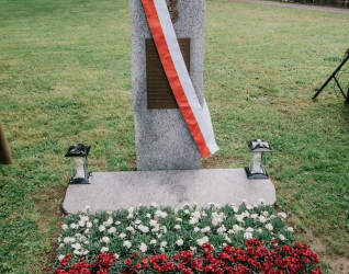 Granitowy obelisk z brązową tablicą i polskim orłem, przewiązany biało-czerwoną wstęgą, otoczony czerwonymi i białymi kwiatami na cmentarzu Madretsch.